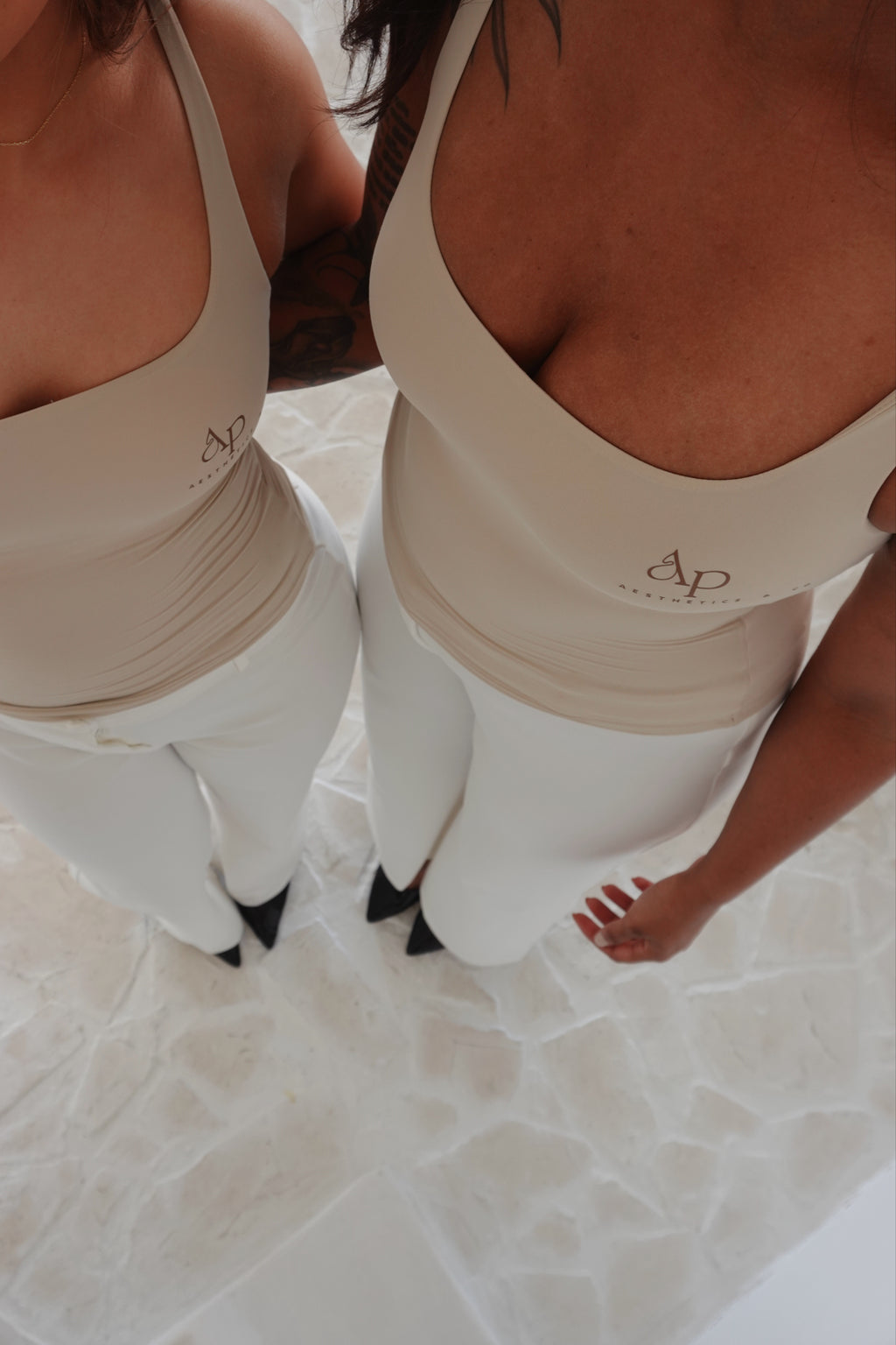 Two women wearing matching beige tank tops with a logo, standing on a light-colored floor.