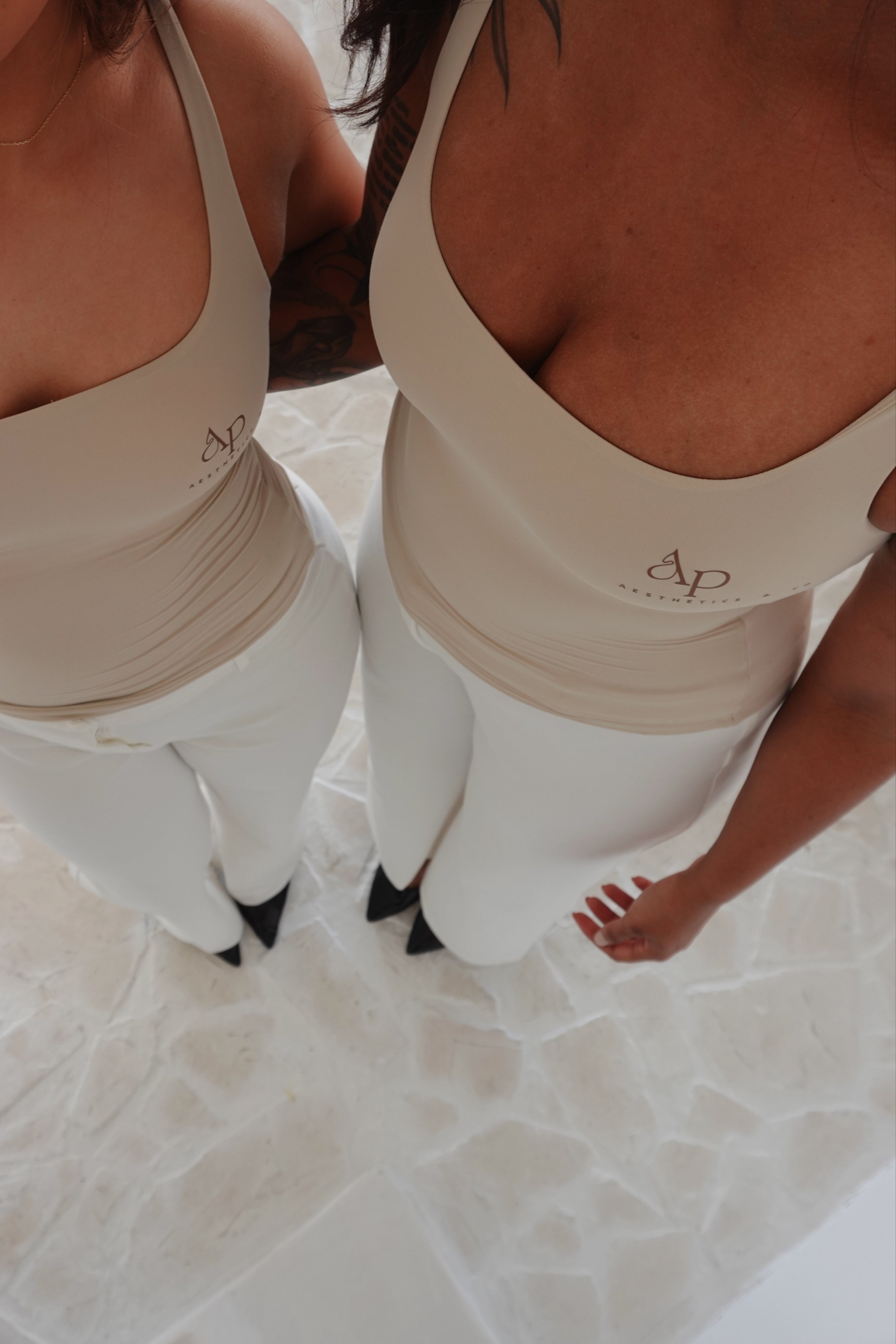 Two women wearing matching beige tank tops with a logo, standing on a light-colored floor.